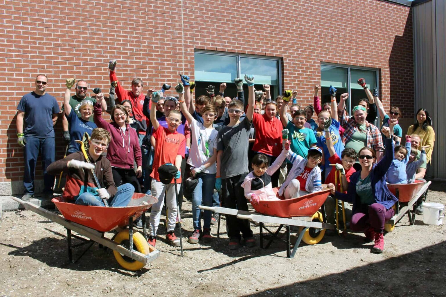 A group of grade school children and teachers wearing work gloves and surrounded with wheelbarrows who are all holding their arms in the air which a sense of accomplishment