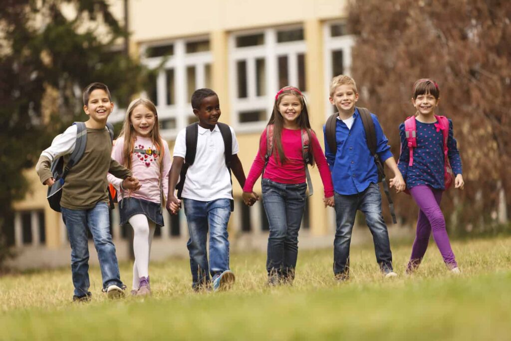 A group of diverse school kids carrying backpacks and holding hand while walking in a line on a grassy lawn