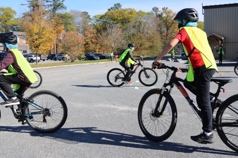 Three grade school-aged kids wearing safety vests and bike helmets maneuvering around small pylons in a school yard