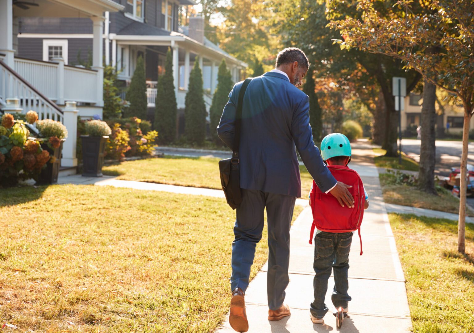 A man patting the back of a young boy on a scooter in a fatherly manner, while they move down the sidewalk of a residential street