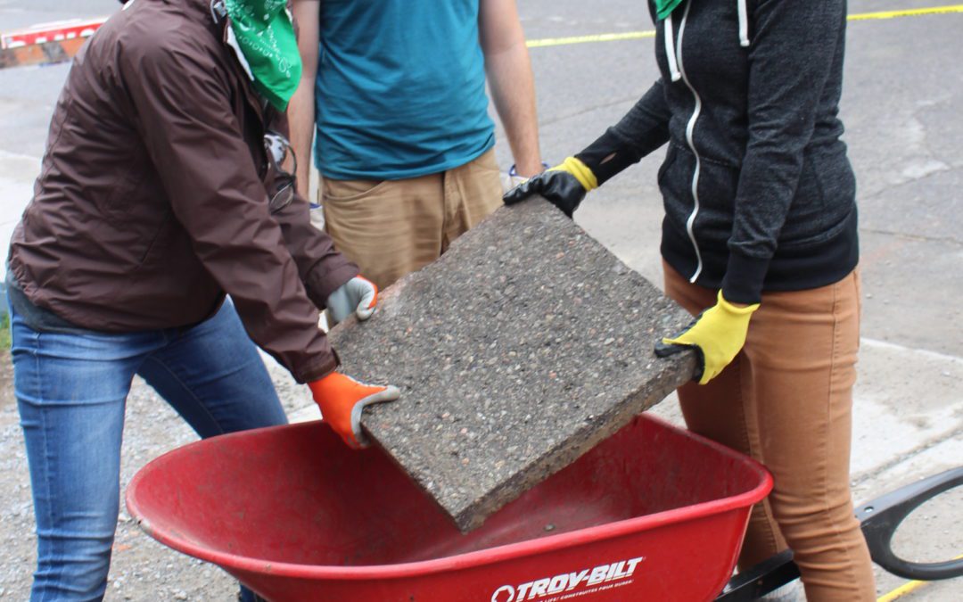 GCC Executive Director Brianna Salmon, GreenUP Depave Coordinator Dylan Radcliffe, and volunteer lift a slab of concrete out of a wheelbarrow.