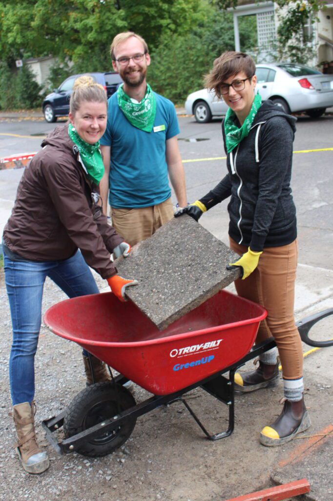 GCC Executive Director Brianna Salmon, GreenUP Depave Coordinator Dylan Radcliffe, and volunteer lift a slab of concrete out of a wheelbarrow.