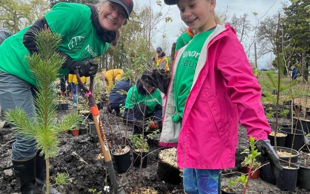 A woman wearing a green t-shirt and baseball hat, and holding a shovel, and a child wearing a pink coat, green t-shirt, and baseball hat, planting trees