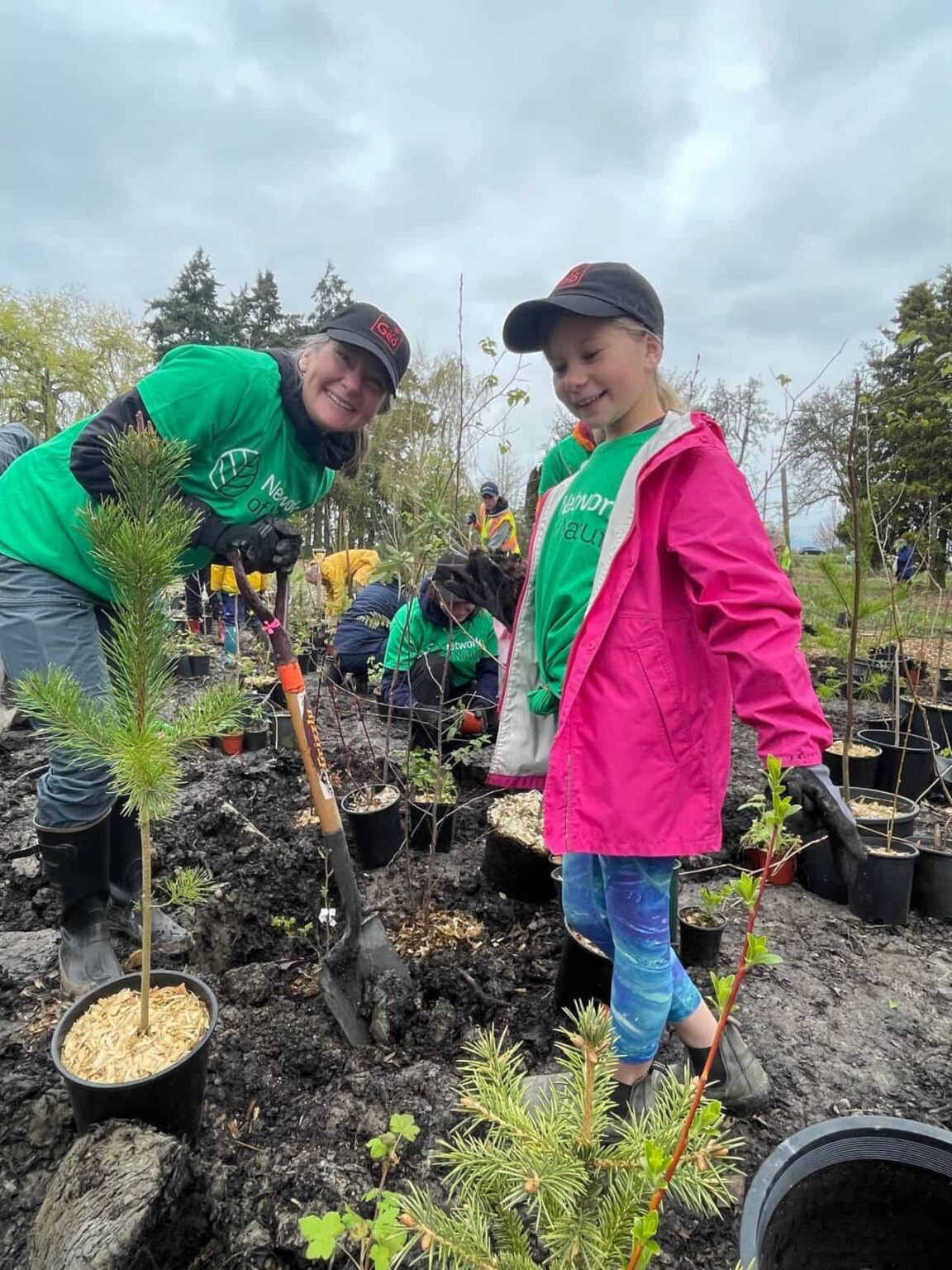 A woman wearing a green t-shirt and baseball hat, and holding a shovel, and a child wearing a pink coat, green t-shirt, and baseball hat, planting trees
