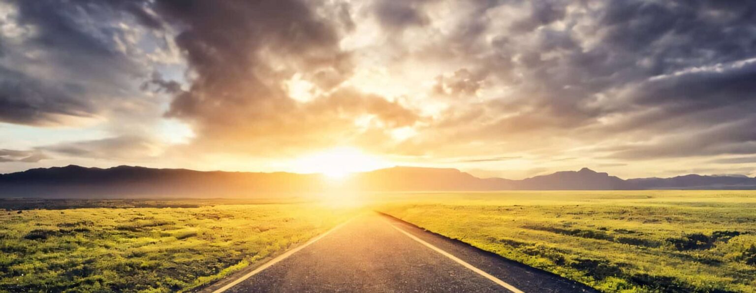 A road leading to the mountains, boarded by green fields.