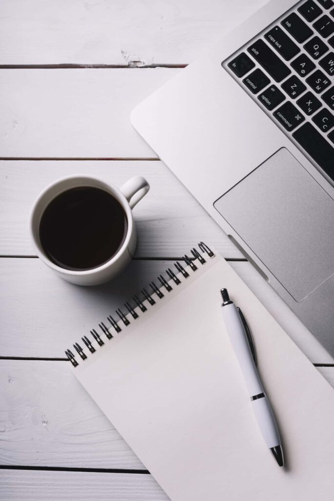 Coffee cup, notepad and pen, laptop on a desk.