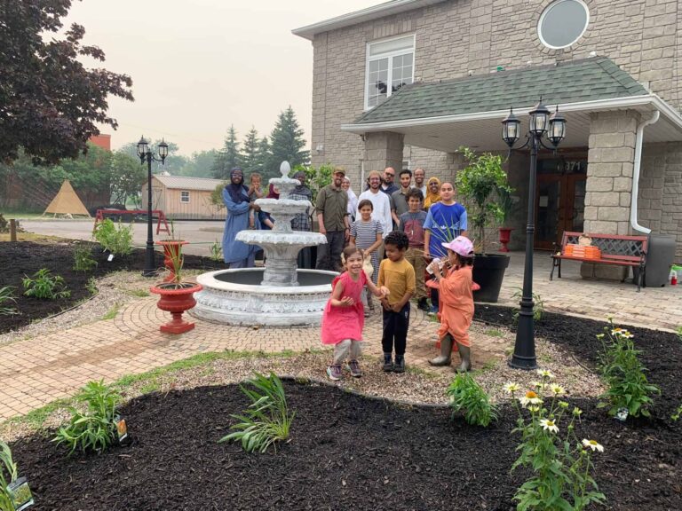 Men, women, and children pose in a straight line in front of the community mosque. The rain garden is in front of them.