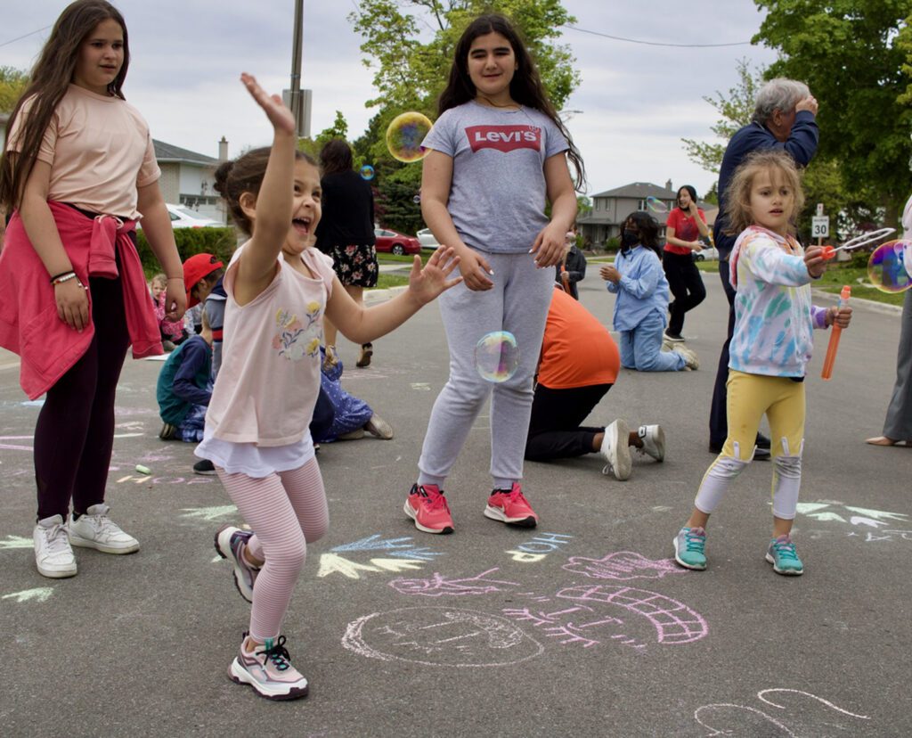 Kids playing on pavement covered in sidewalk chalk reaching for bubbles, drawing on the pavement or interacting with each other with smiles on their faces