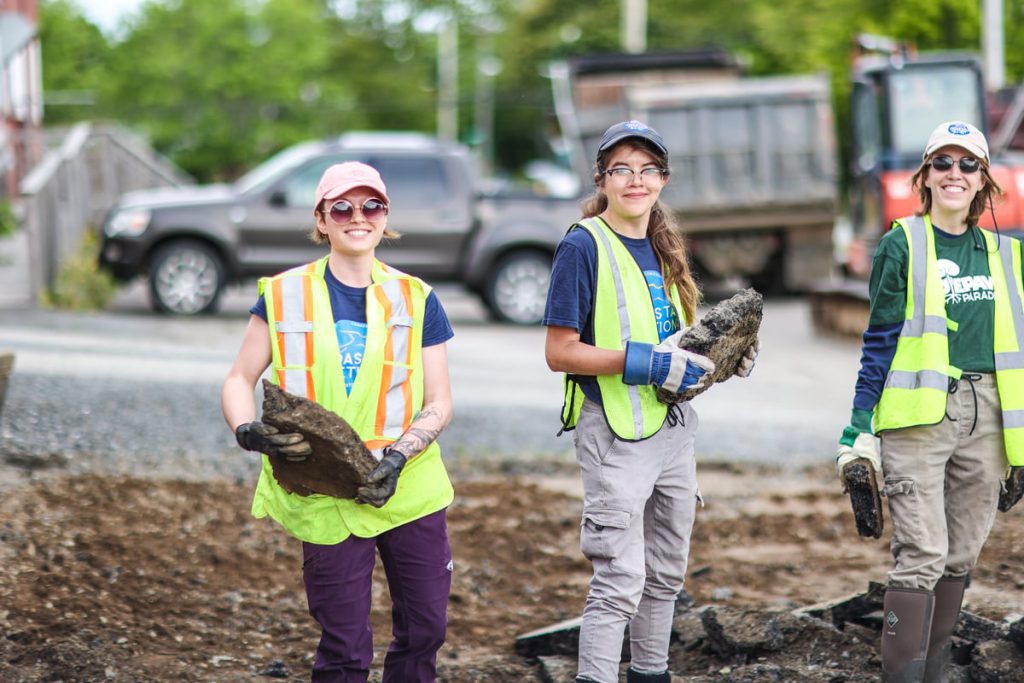 A group of smiling volunteers outdoors in front of a building, wearing bright yellow safety vests and gloves, working together to remove pavement.