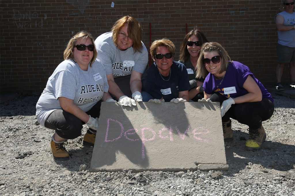 Five smiling adults crouch together outdoors, holding a slab of asphalt with the word “Depave” written on it in pink chalk. They wear work gloves and sunglasses, and some wear shirts that say 'Rideau Heights'.
