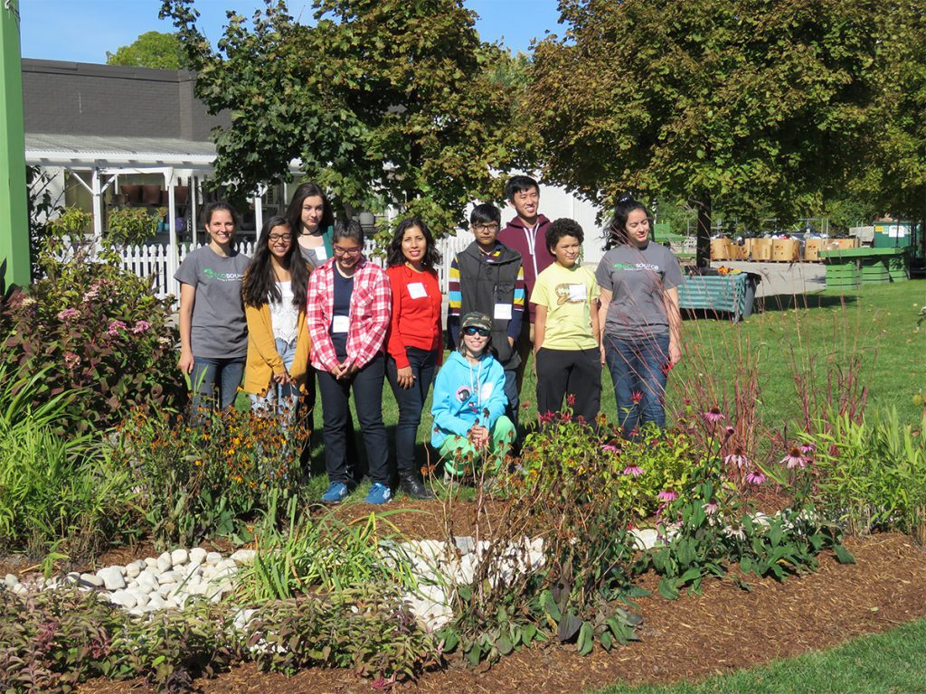 A group of ten people stand in a field behind a rain garden filled with flowers and other plants. They’re smiling at the camera, with a plant nursery and trees visible in the background.