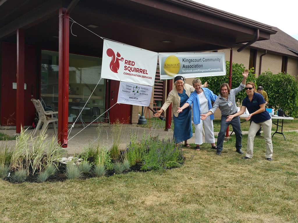 Four people stand on grass next to Kingscourt Free Methodist Church, posing with their arms open wide beside a newly installed rain garden.