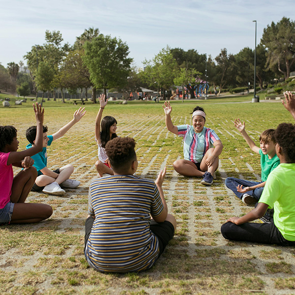 Seven students sitting in a circle, in a grassy park with their hands raised in the air as if to ask a question