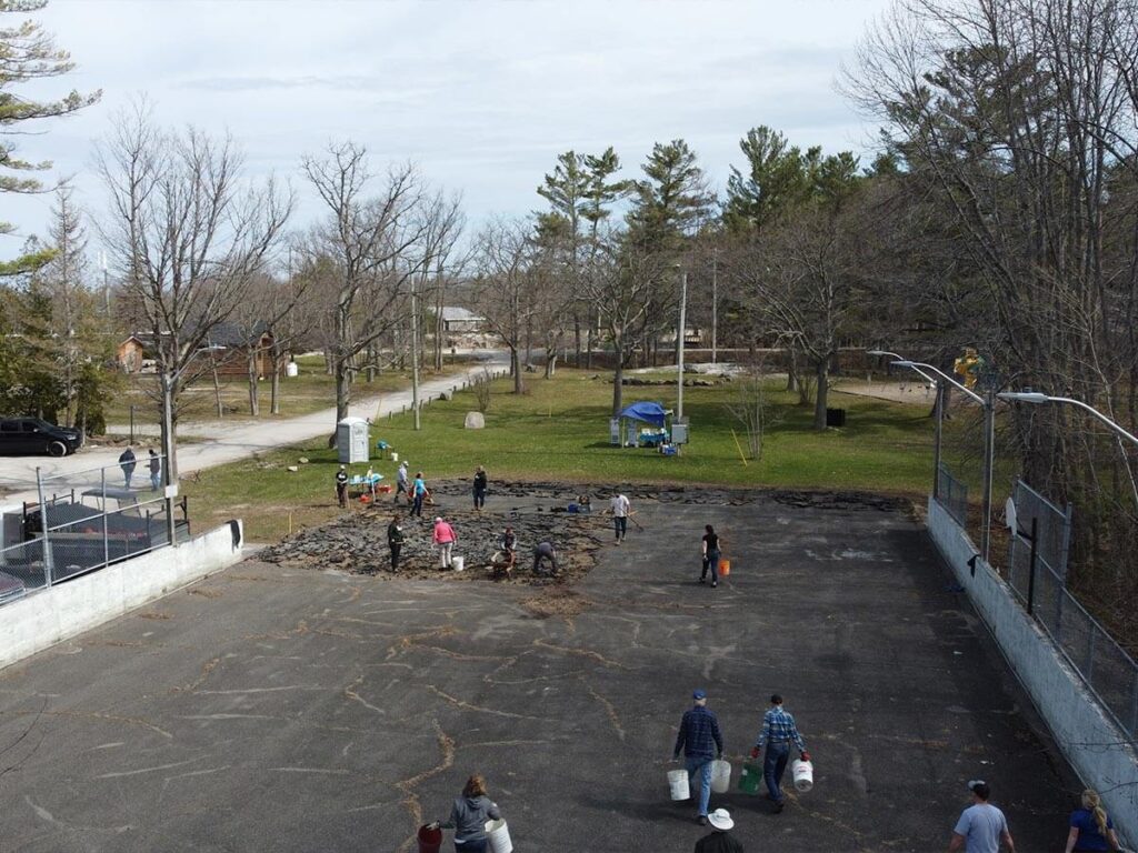 Aerial photo of volunteers depaving an unused skating rink, surrounded by grass, trees, and a parking lot on one side. Over fifteen people carry buckets and use tools to remove sections of pavement.