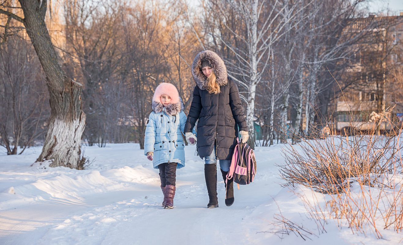 An adult and a child walk hand in hand through snow, wearing winter coats with fur-lined hoods.