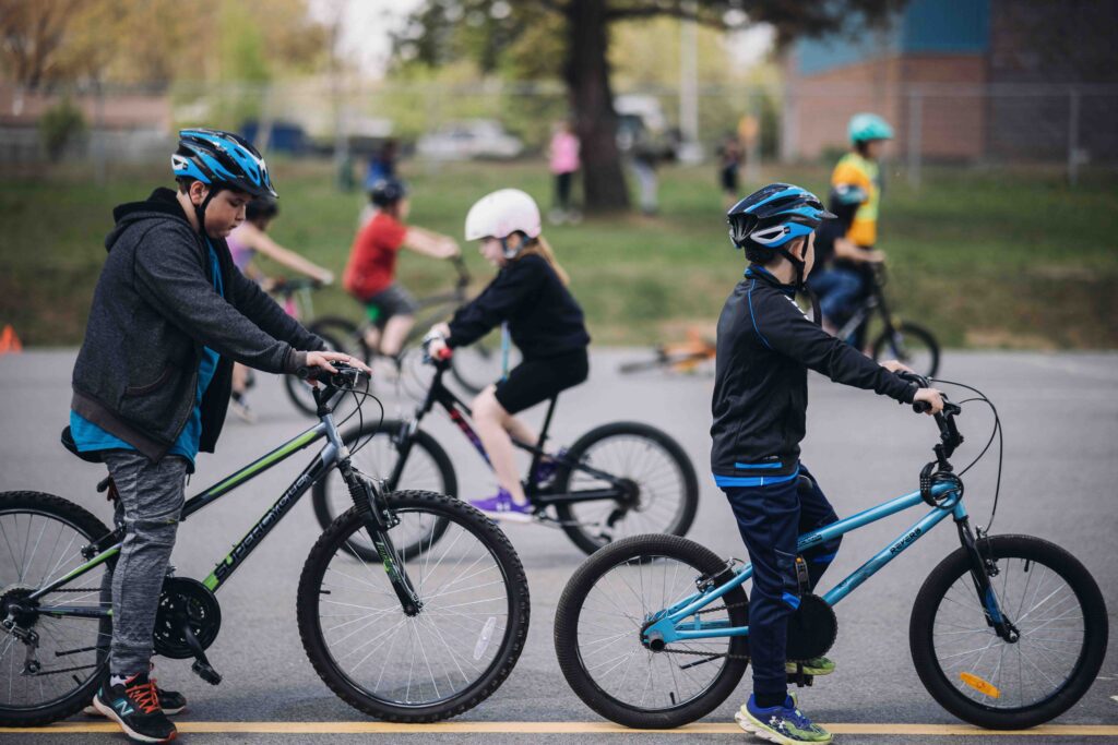 Two kids on bikes waiting on a painted line for their turn in an bike skills course. Blurred background shows other kids completing the skills course.