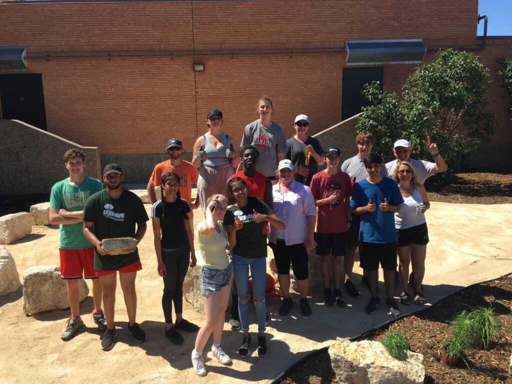 A group of sixteen students and adults smiling in front of a newly depaved pathway surrounded by mulch at Miles Macdonell Collegiate.