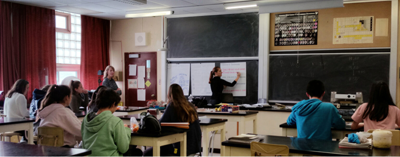 Students seated in a classroom listening to a teacher speak.