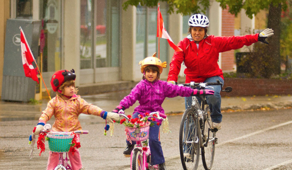 A bike train with two children and an adult riding bicycles together, all wearing helmets and extending their arms to signal.