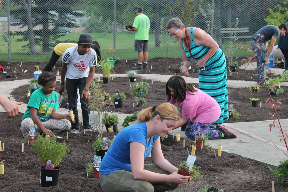 A group of people planting in a community garden with freshly turned soil. Small plants in pots and markers show the planning of the garden.