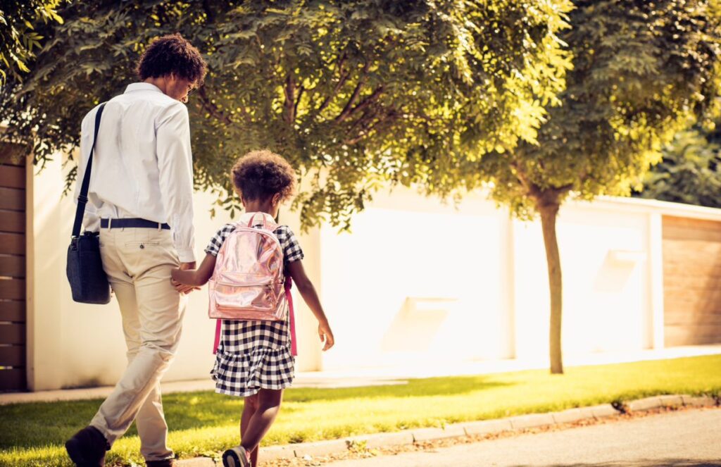 An adult and a child walk hand in hand along a tree-lined street, with the child wearing a backpack.