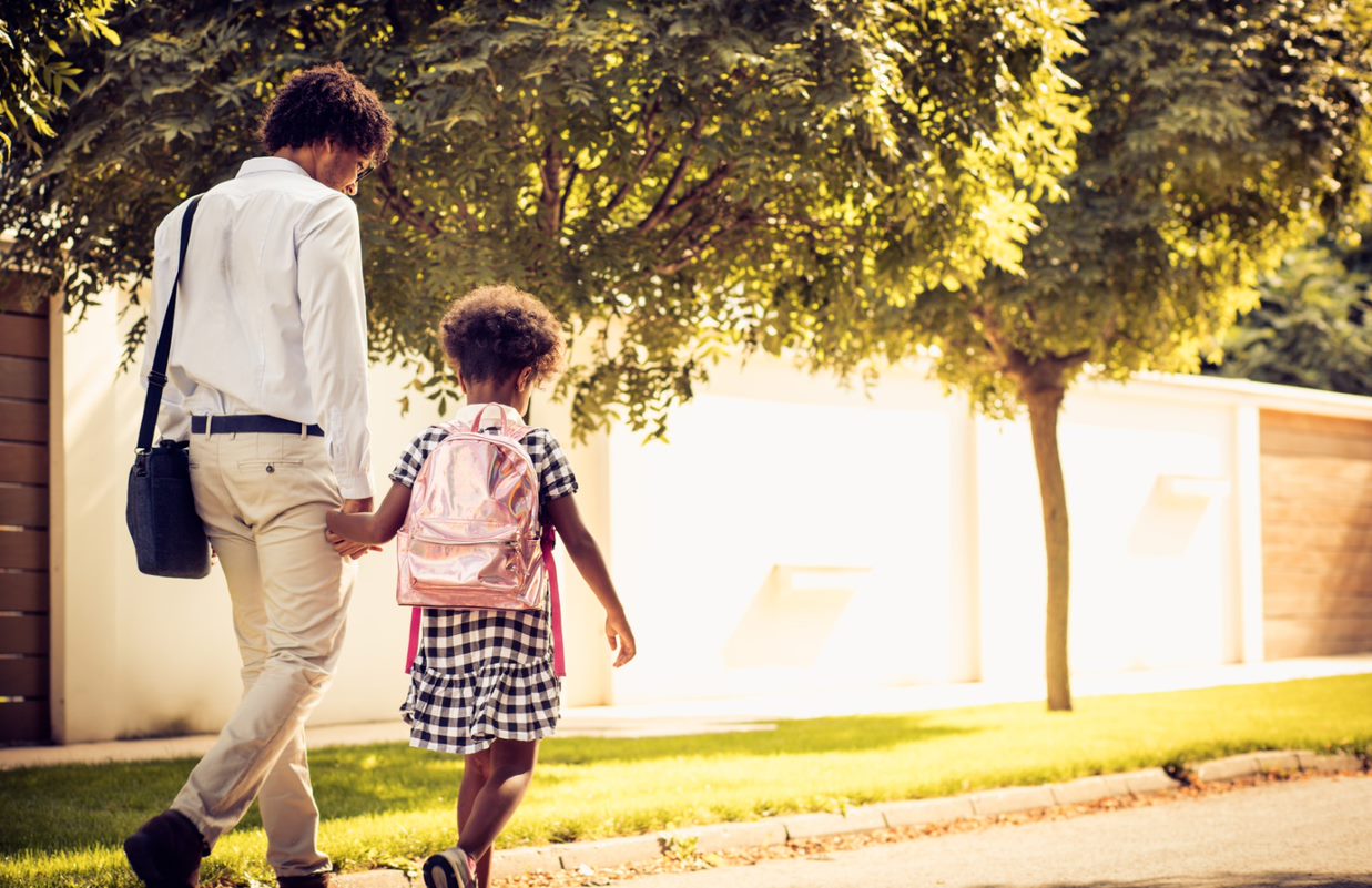 An adult and a child walk hand in hand along a tree-lined street, with the child wearing a backpack.