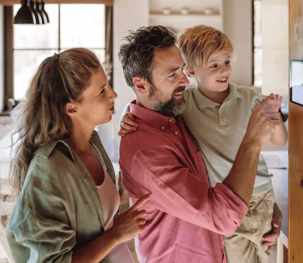A family changing settings on their home thermostat, the father carries the child in his arms.