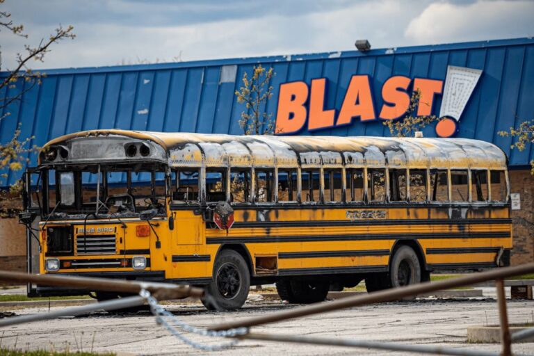 Photo of burnt yellow school bus with sign in background that says "BLAST"