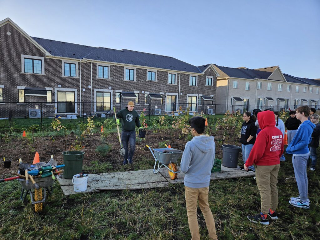 Students stand around a man holding a shovel. He is providing a planting tutorial with various plants and equipment surrounding him. A row of the backside of townhomes can be seen in the background.