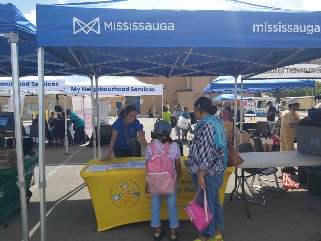 A city of Mississauga event tent with staff person inside smiling and talking to a parent and child.