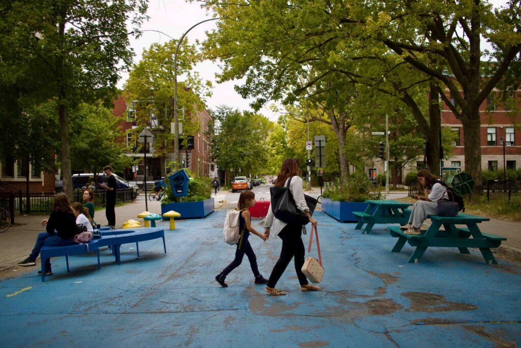 Families using a painted pedestrianized street with picnic and other benches, with trees overhead.