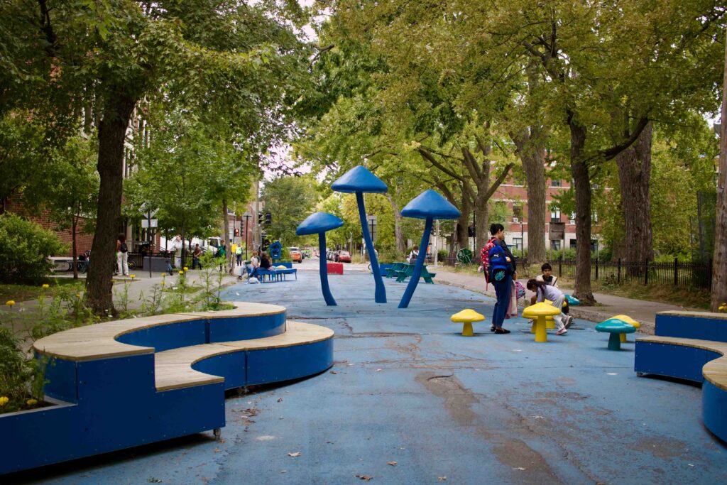 Pedestrianized street with large mushroom urban art sculptures, overhanging trees, and benches, with kids and families enjoying the space.