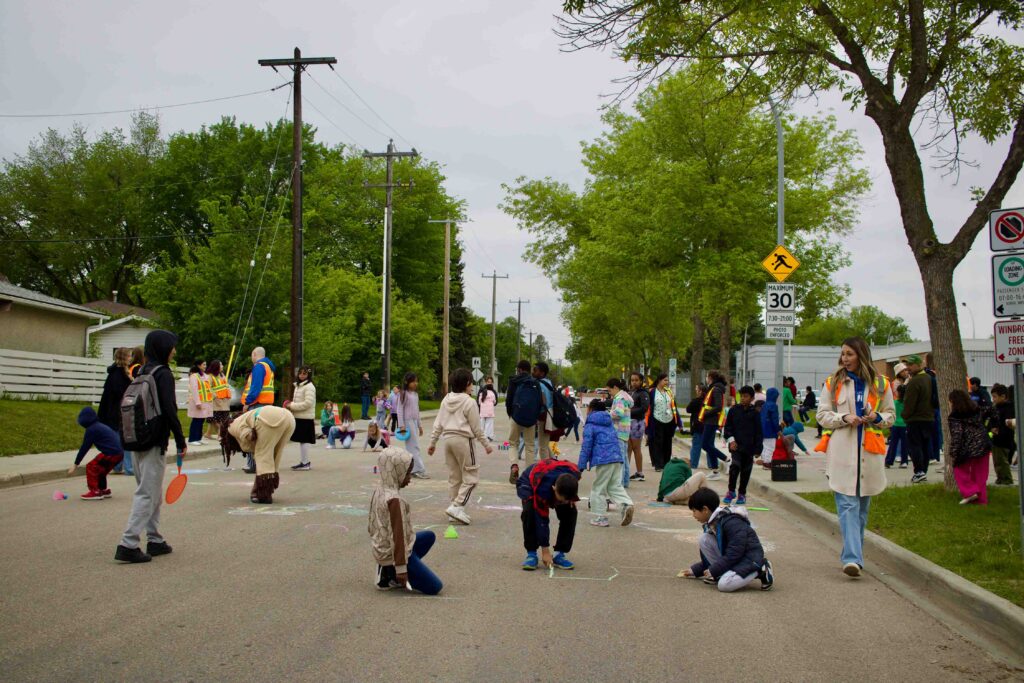 Dozens of children drawing chalk and playing games on a pedestrianized street in front of a school.