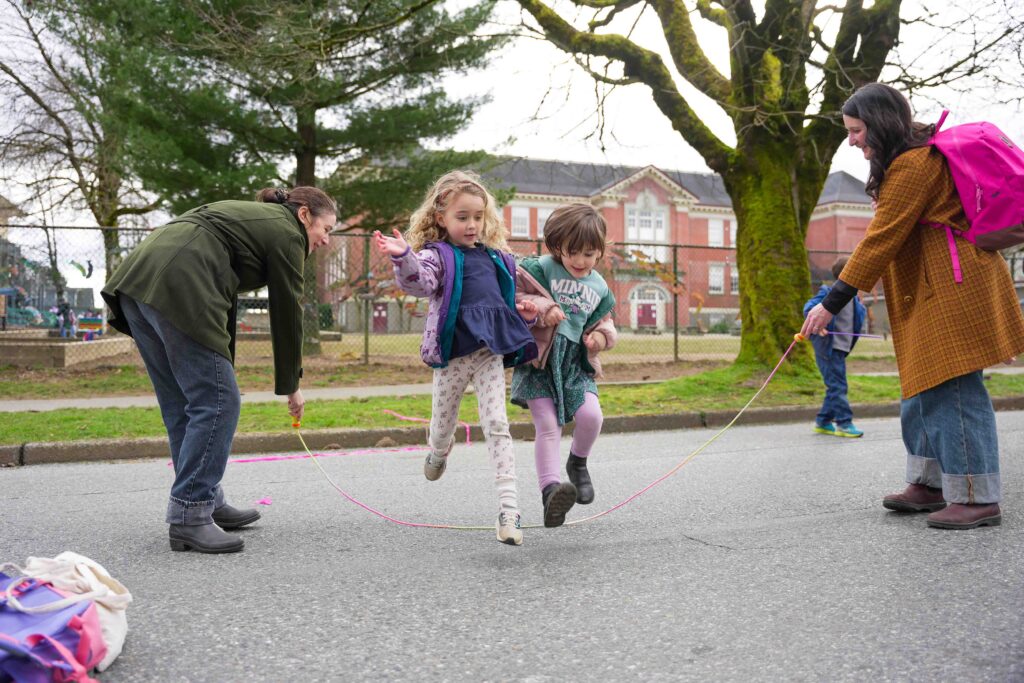 Two children leap over a jump rope swung by two adults on a pedestrianized street in front of their school.