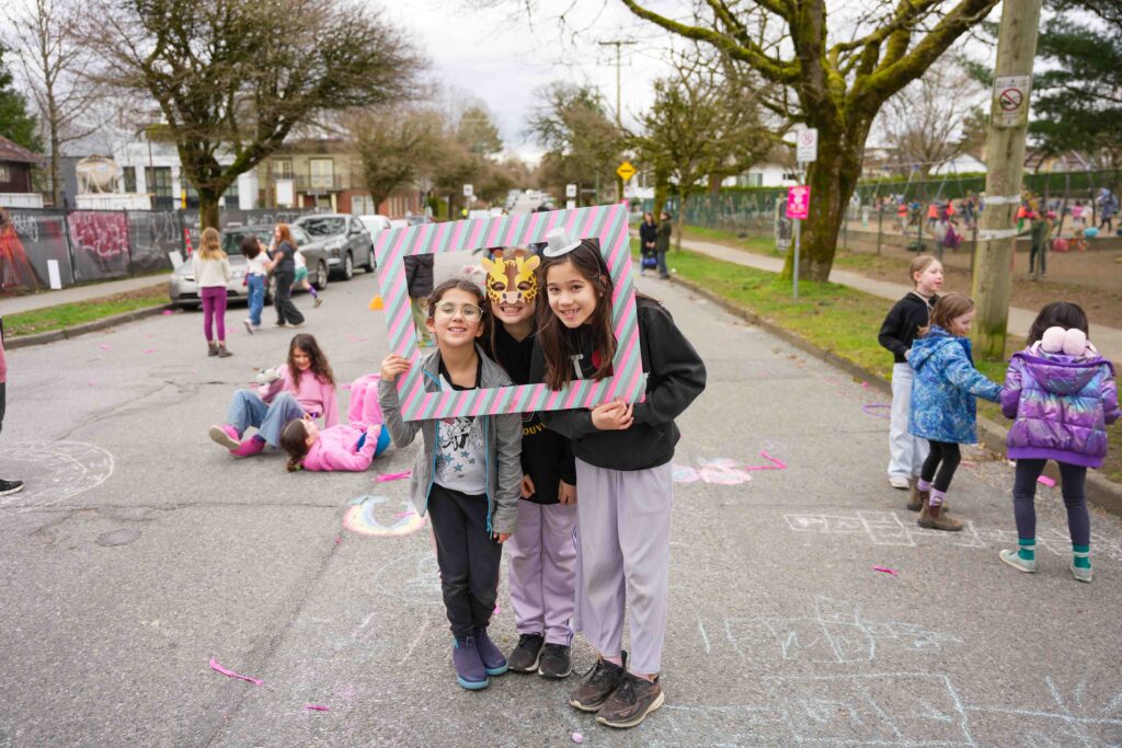 Three kids with masks and hats smiling as they hold a colourful frame around their faces. In background other kids play on a pedestrianized street in front of a school.