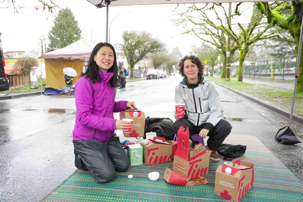 Two adults preparing to hand out hot chocolate on a rainy day street in front of a school.