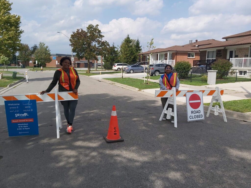 Two people wearing safety vests and managing temporary street barriers with multi-lingual school streets signage. They smile at the camera