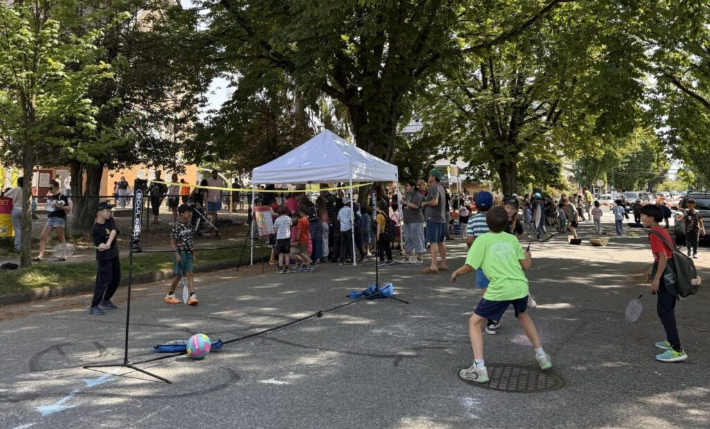 Kids playing badminton on a pedestrianized street in front of a school. In background, an event tent with many other kids gathered around and playing on the street.
