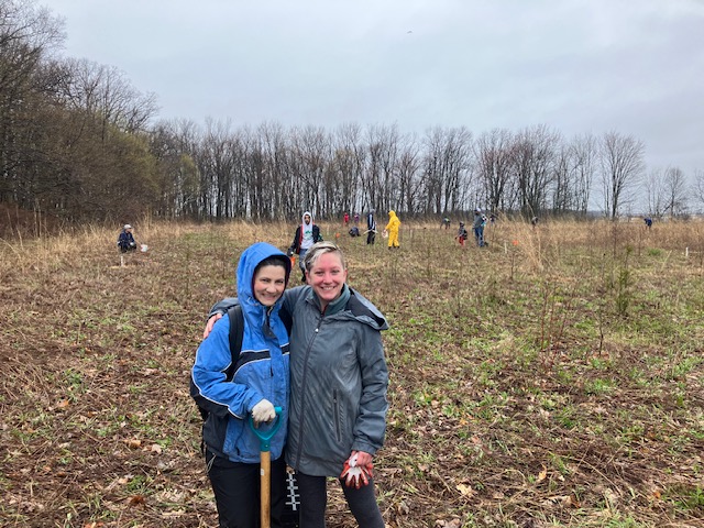 Two people smile. A former farm field is visible behind them with more people surveying and planning to reforest the space. 